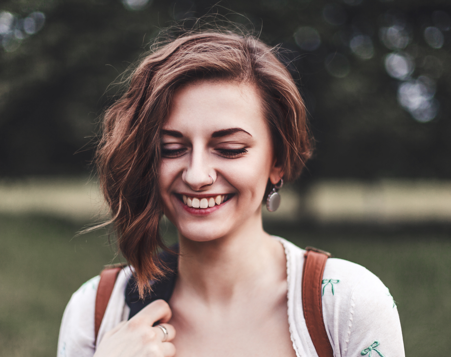 Retrato de uma mulher que está sorrindo e olhando para baixo. Ela está na rua, em um ambiente bem solar.